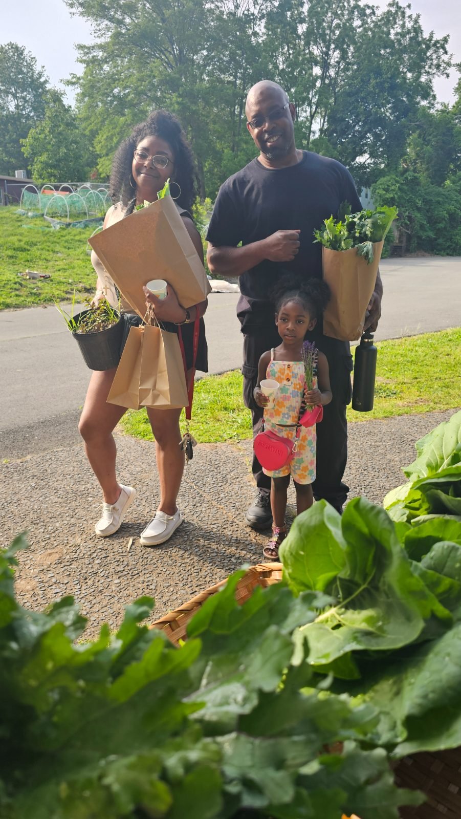 A family at pickup — two adults and a small girl with paper bags of greens, a pot of seedlings and a bunch of lavender.