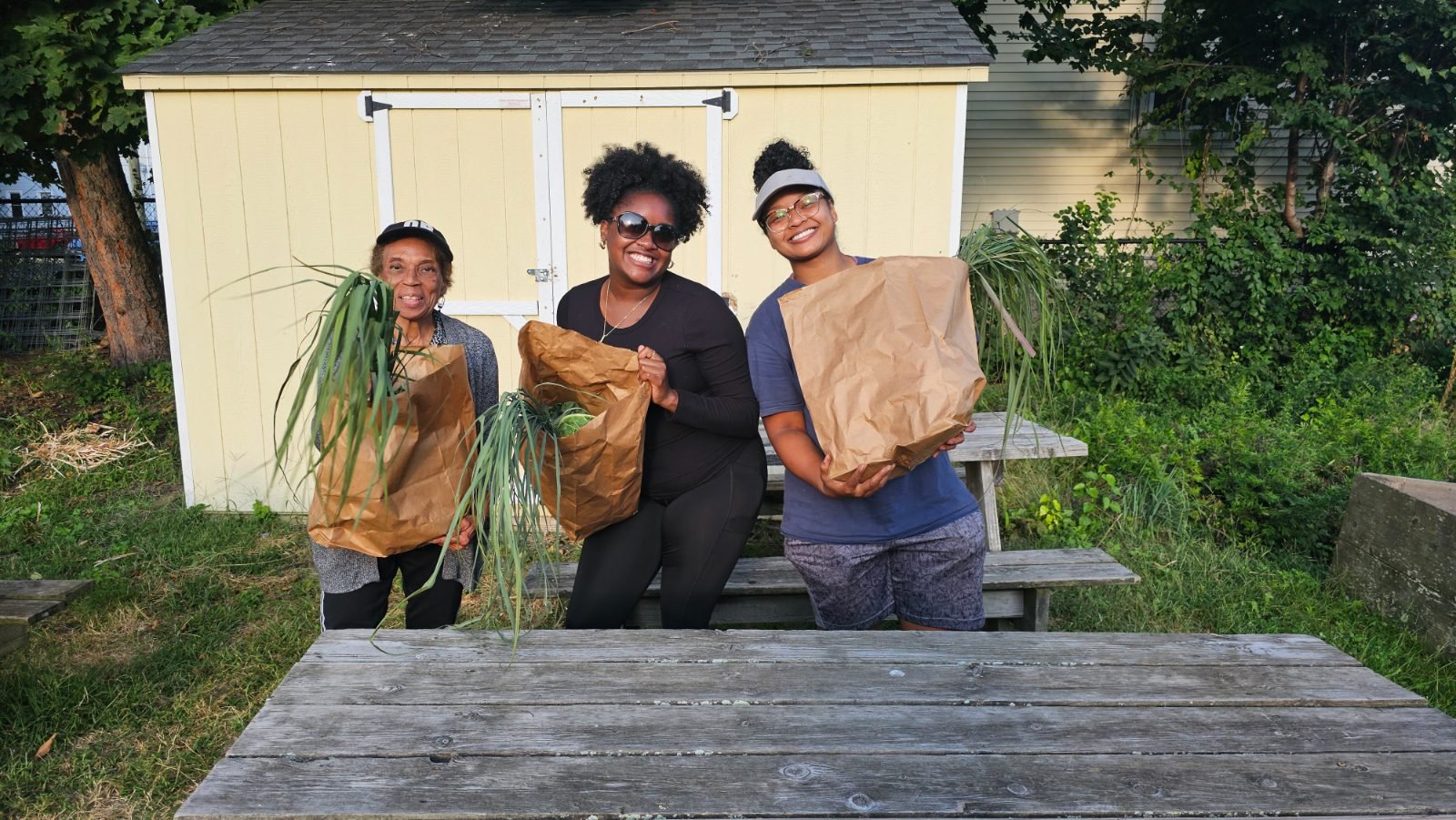 Three generations behind a picnic table, each holding a paper bag with the tops of freshly pulled alliums spilling out.