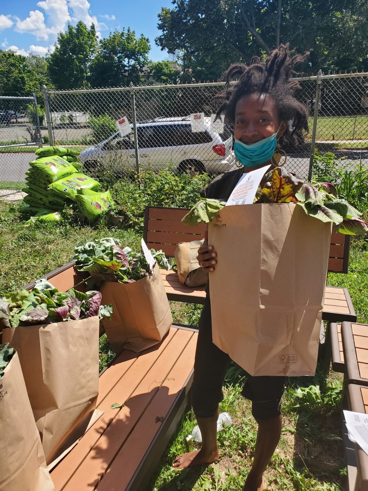 A neighbor holding up a full kraft sack of amaranth and collard on pickup day, more packed sacks waiting on the bench beside her and the garden beds behind.