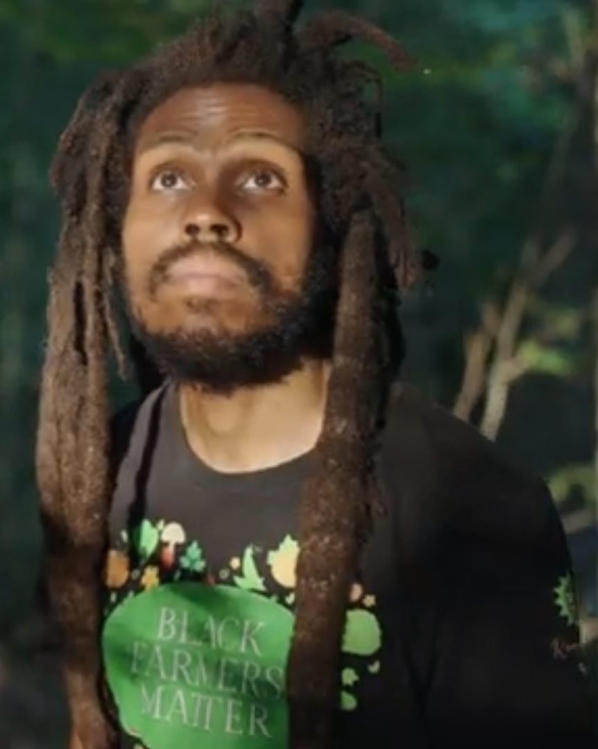 A close portrait of Farmer D in a &ldquo;Black Farmers Matter&rdquo; apron, greenery behind.