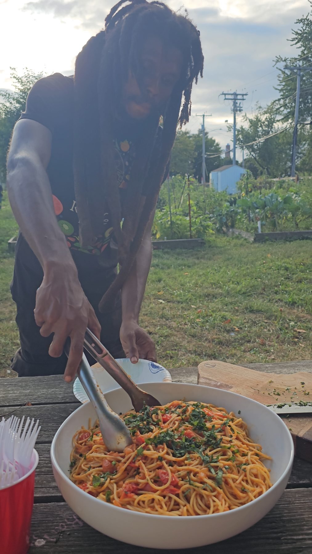 Hands serving spaghetti with fresh tomato sauce and chopped herbs from a large bowl on a garden picnic table.