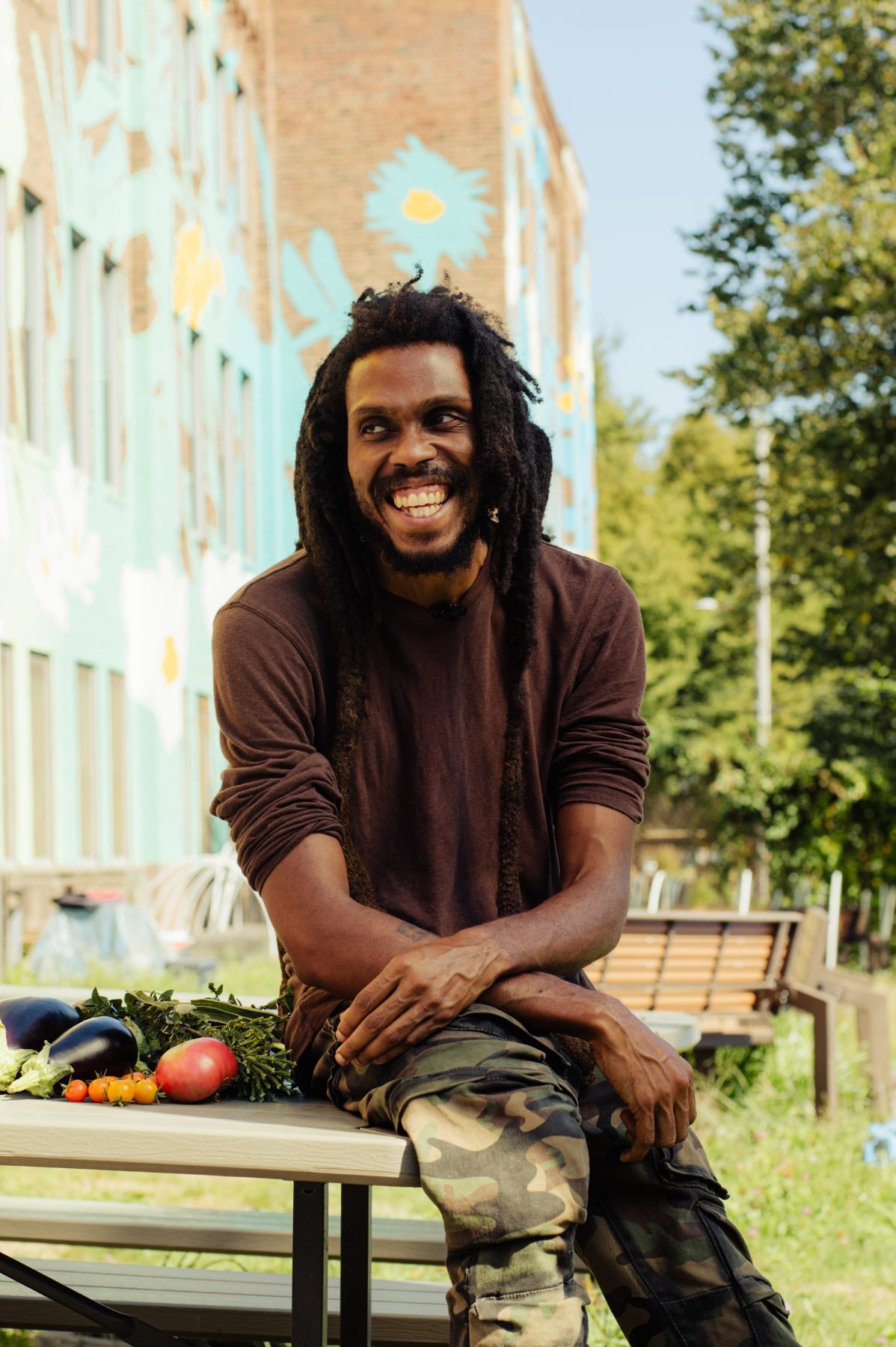 A farmer sitting on a garden picnic table beside eggplant, tomatoes and herbs, laughing, a painted mural wall behind him.